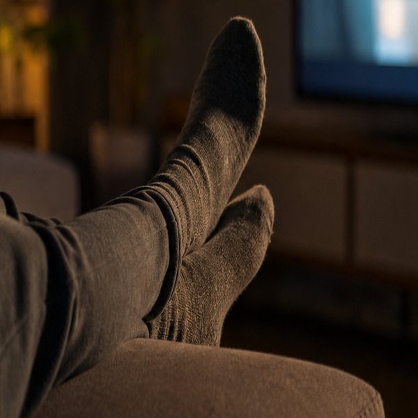 A person sits on a couch in dim lighting, holding a remote control while watching a television screen. — detail